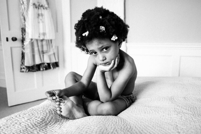 Photo portrait of a flowergirl sitting on a bed. She is resting her head on one hand and with the other hand she is playing with a hairband.