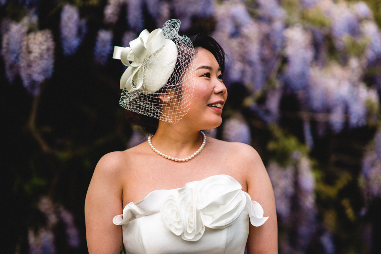 A bride at the Graduate Cambridge. She is wearing a small white hat and wisteria is behind her.