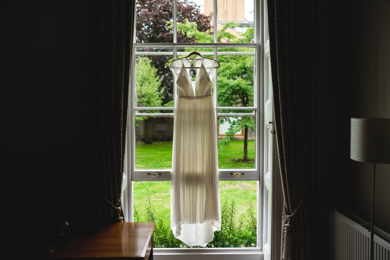 A wedding dress hangs on a window. Behind is the grounds of Downing College.