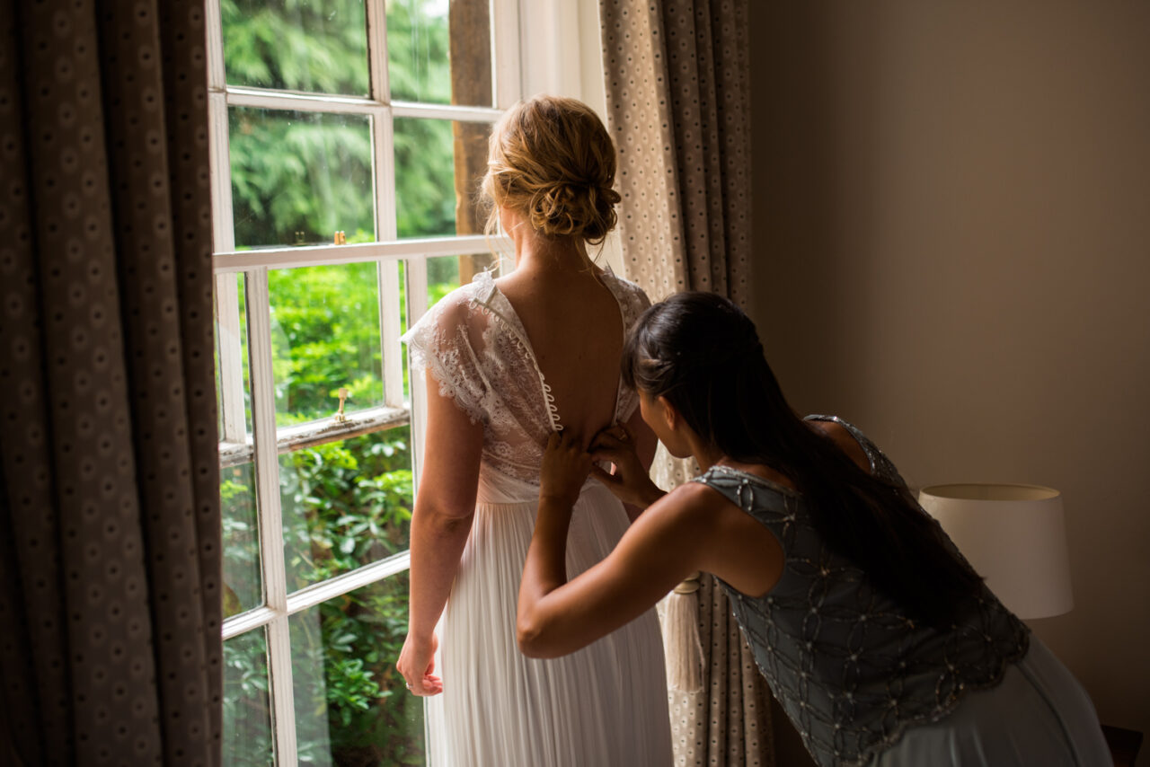 A bridesmaid adjust a bride's dress by a window at Downing College.