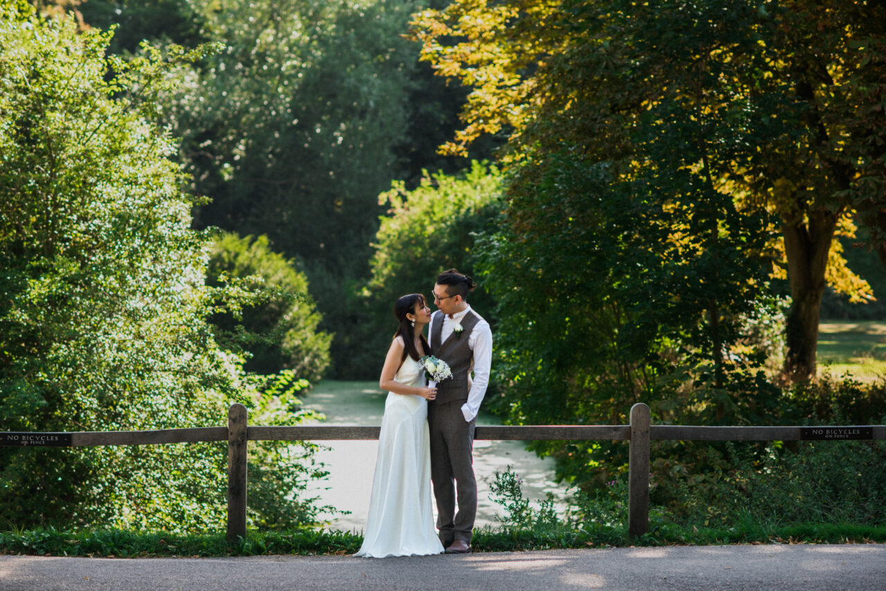 A couple pose in The Backs in Cambridge with a stream and many trees behind them.