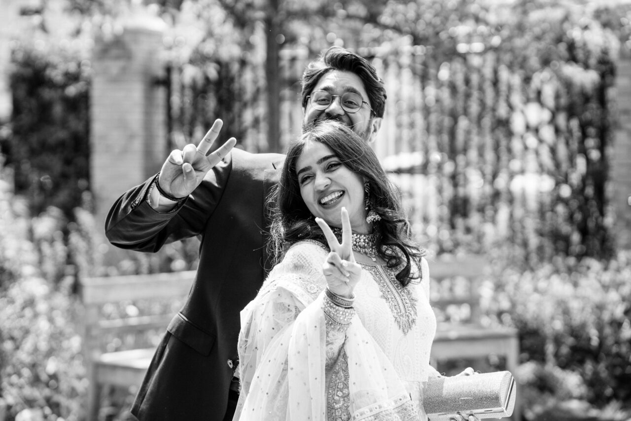 A bride and groom pose with their fingers making a V sign in the gardens of Cambridge Central Mosque.