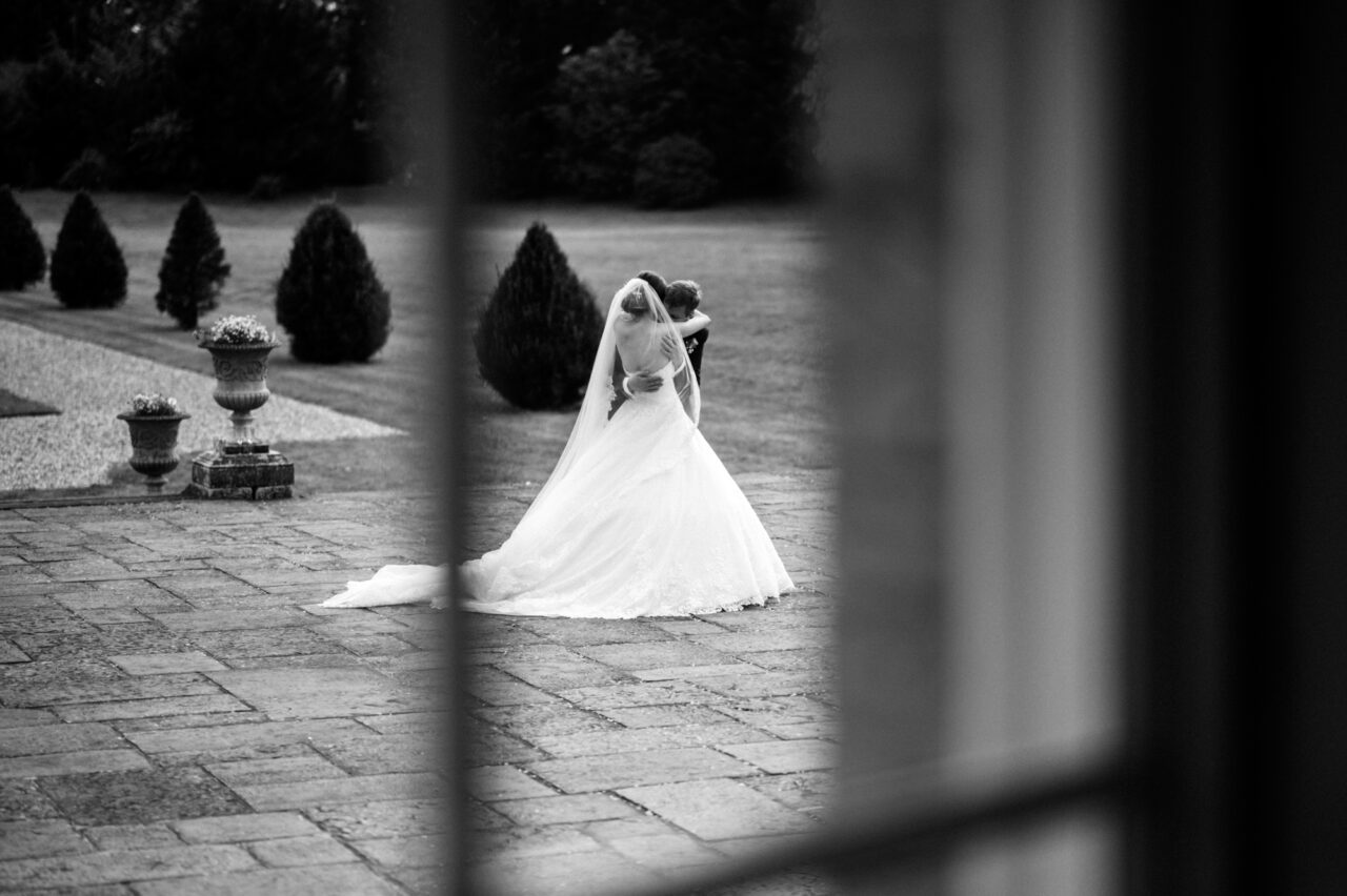 Black and white candid wedding photo of a bride and groom embracing, taken through a window.