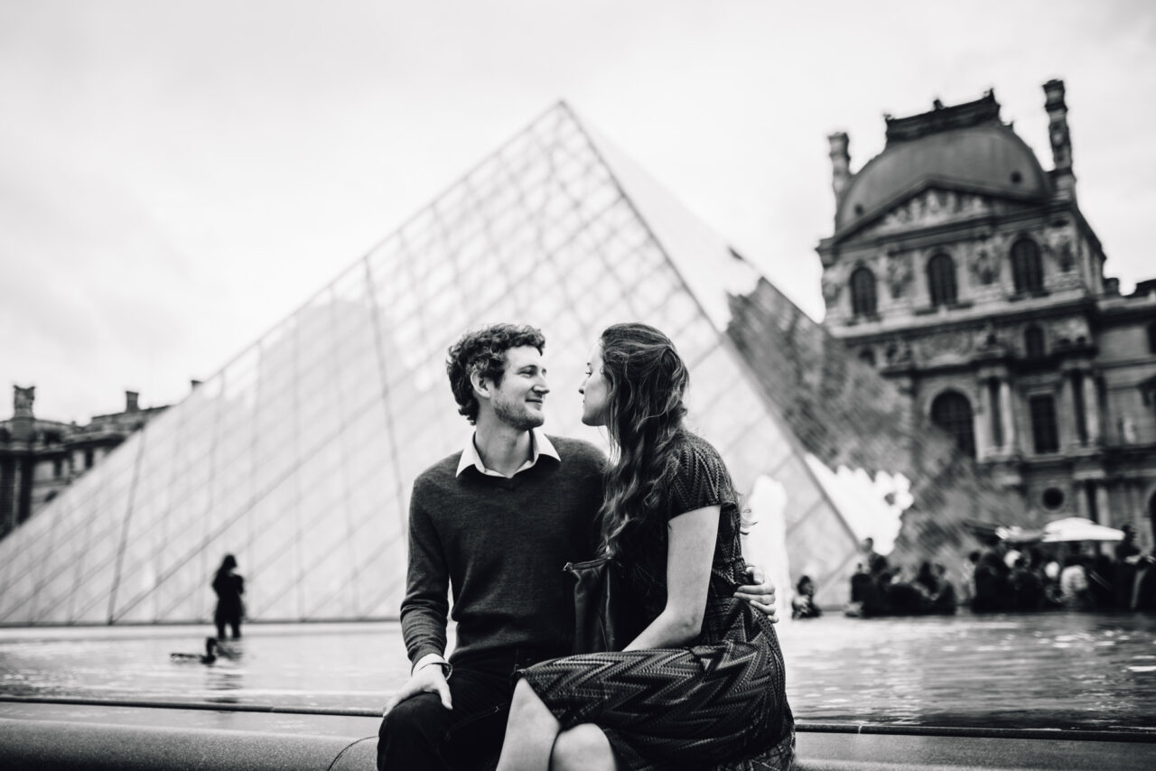 A couple look into each others' eyes sitting on a step near The Louvre in Paris.