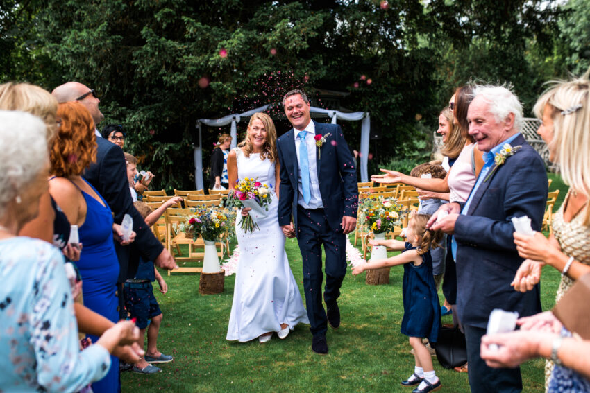 Bride and groom walking through confetti after their ceremony in the gardens at Island Hall wedding venue Cambridgeshire.