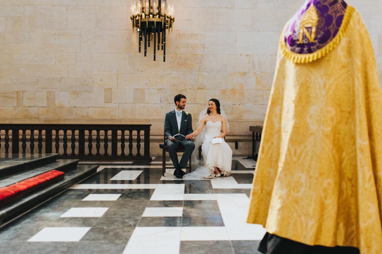 A bride and groom hold hands while seated during their wedding ceremony. In the foreground is the priest's bright yellow robe.