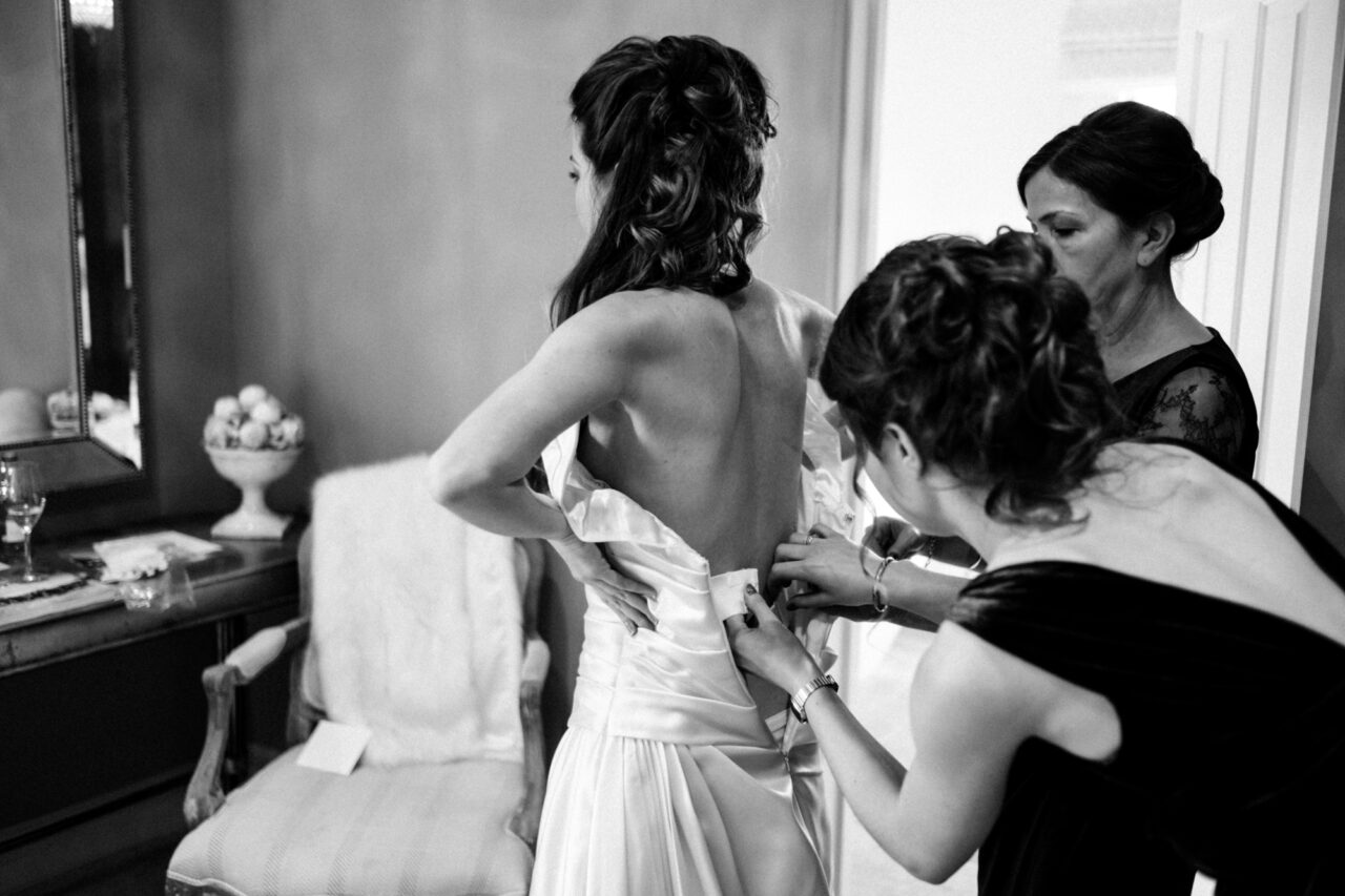 A bride is helped into her wedding dress by her mother and a bridesmaid.