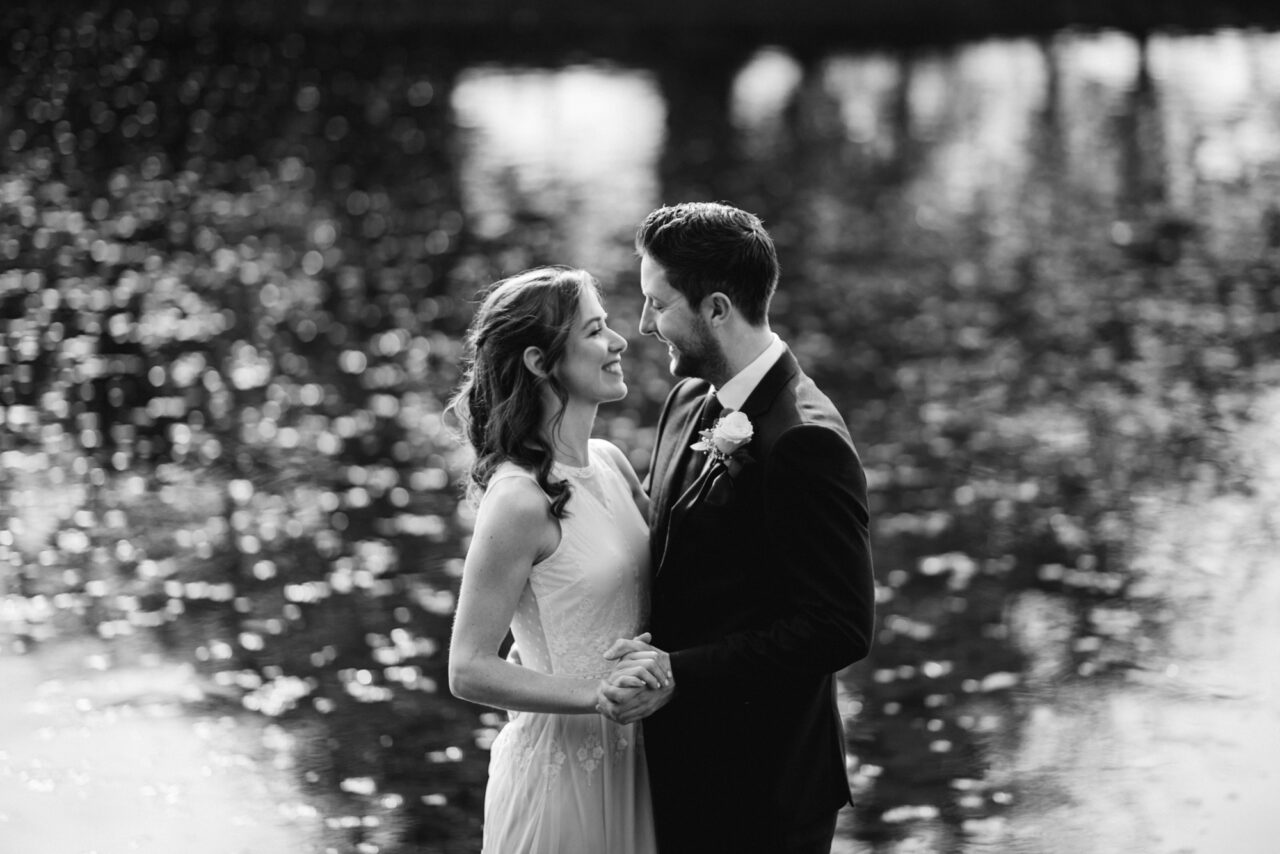 A bride and groom pictured with an out of focus river behind them that is sparking in the sun.