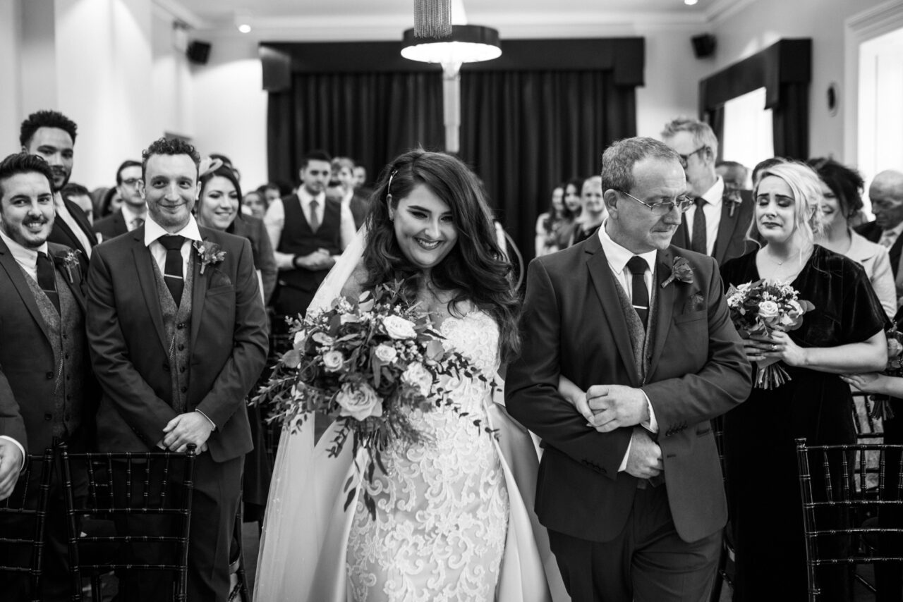 A smiling bride and her father walk down the aisle inside Swynford Manor for her wedding.