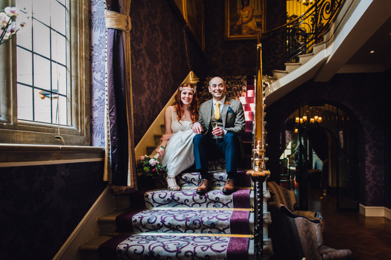 A couple pose smiling on the stairs of their wedding venue.