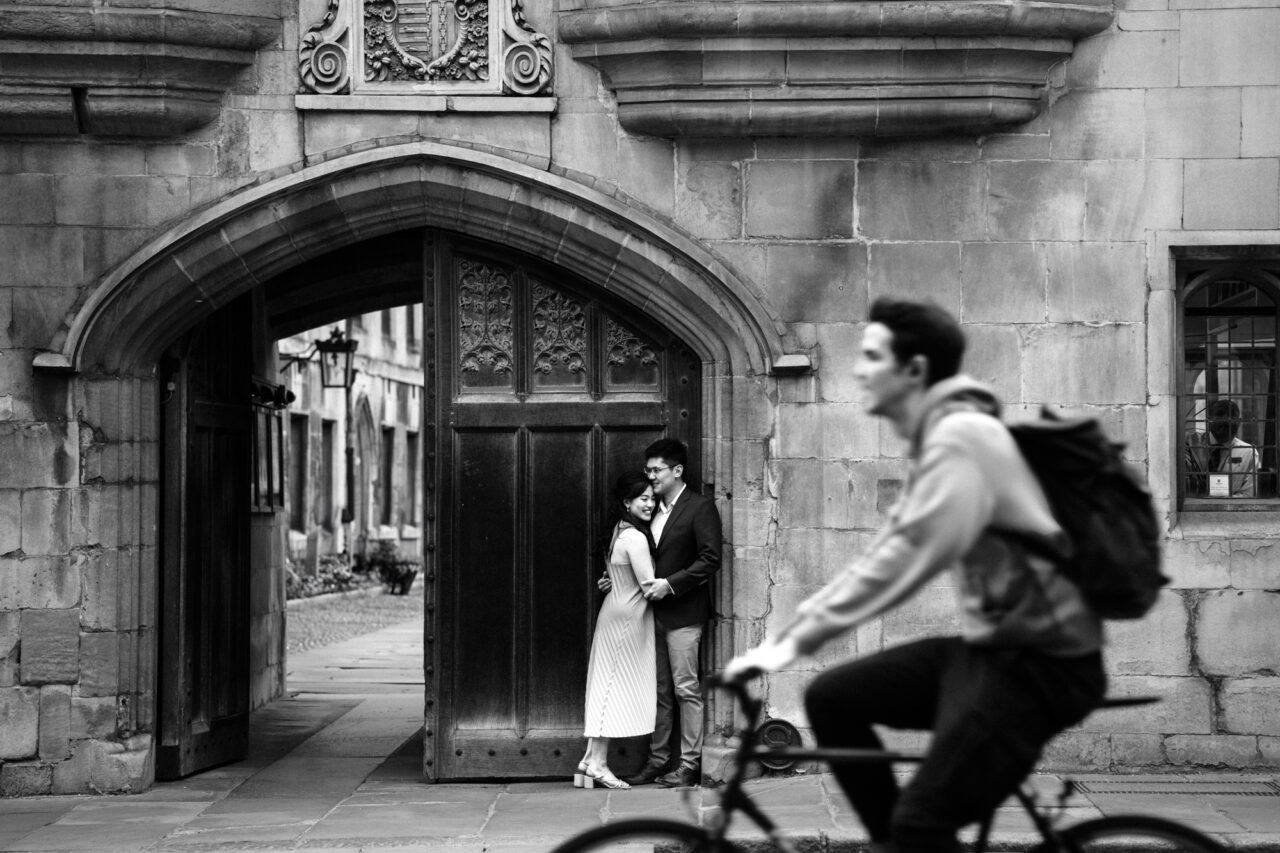 A couple embrace by the entrance to Pembroke College in Cambridge as a cyclist passes by.