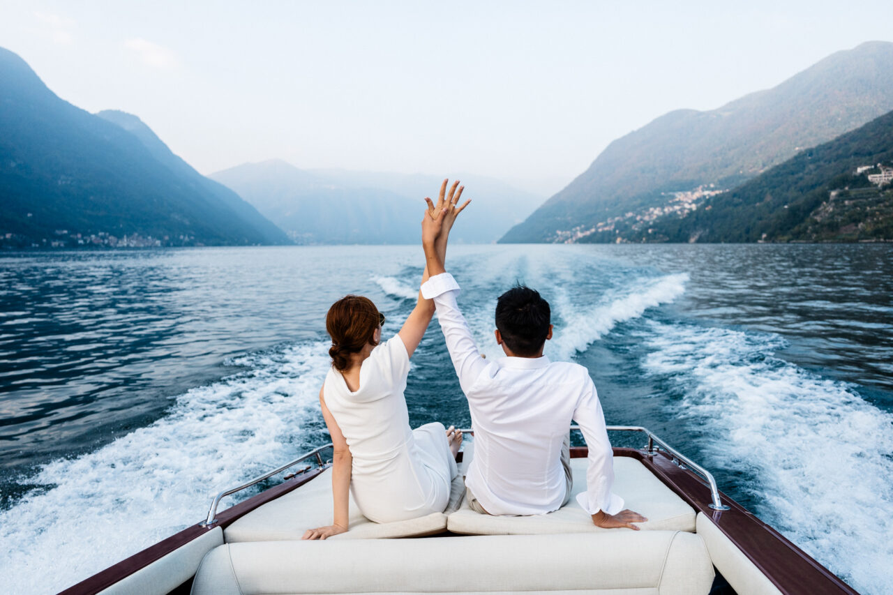 A couple are on a boat in lake Como and are holding their hands up showing an engagement ring.