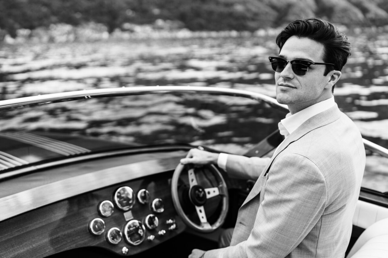 A man poses for a photo while driving a boat on Lake Como. The photo is black and white and the man is dressed very smartly in a suit.