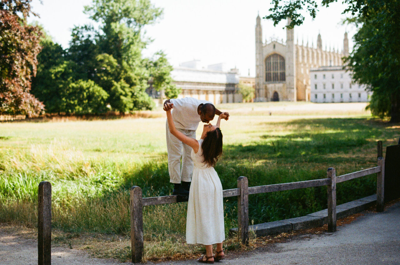 A man stands on a fence kissing his girlfriend at The Backs in Cambridge. Behind them is a field King's College Chapel.