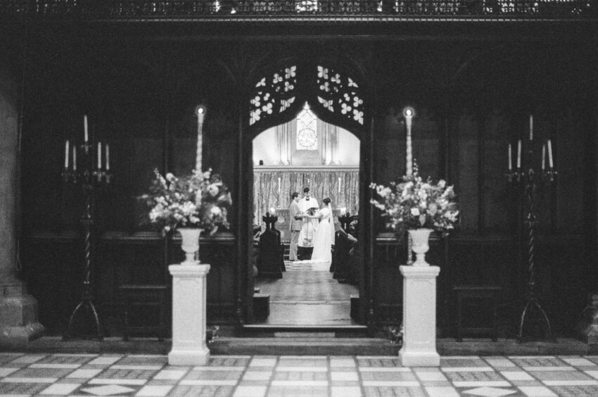 Wedding ceremony at Jesus College in Cambridge photographed on Ilford Delta 3200 film. The bride and groom are holding hands.