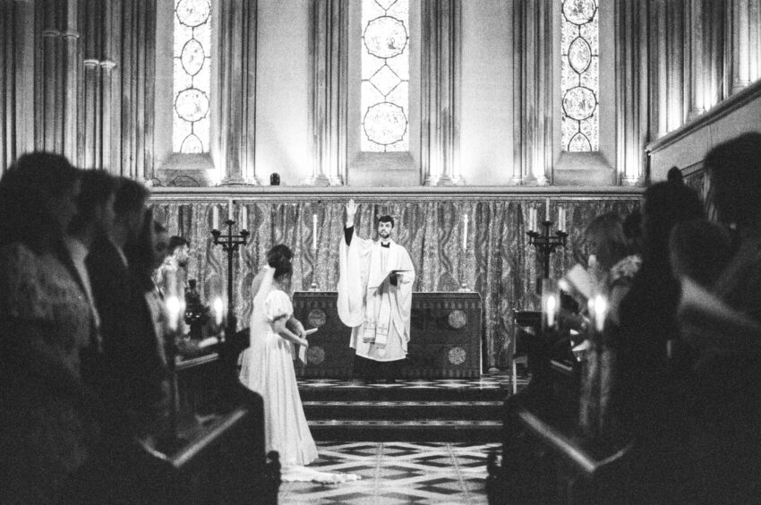 Grainy black and white 35mm film photograph from a wedding ceremony - a priest gives a sermon in a Cambridge chapel.