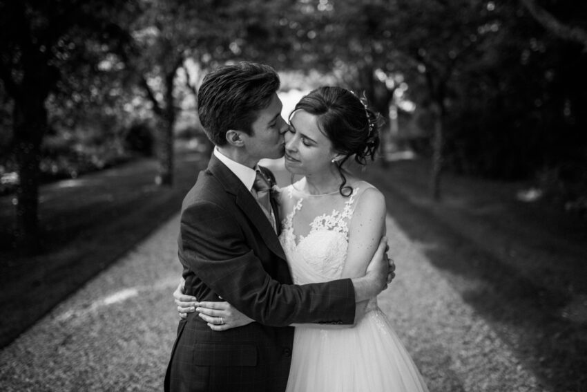 Black and white image of a couple embracing with trees and beautiful light behind them.