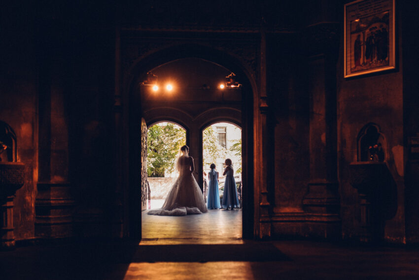 A bride and her flowergirls wait by the entrance of an old church. Light shines in from the open doors.
