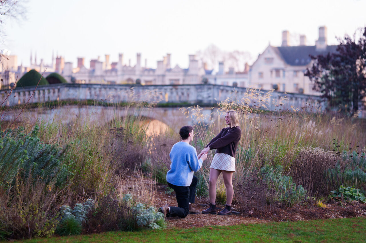 A man proposes to his girlfriend in the gardens of Clare College in Cambridge. The man is on his knees and the lady is smailing.
