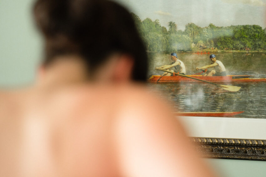 Bride getting ready, softly out of focus, with a rowing scene in a framed picture