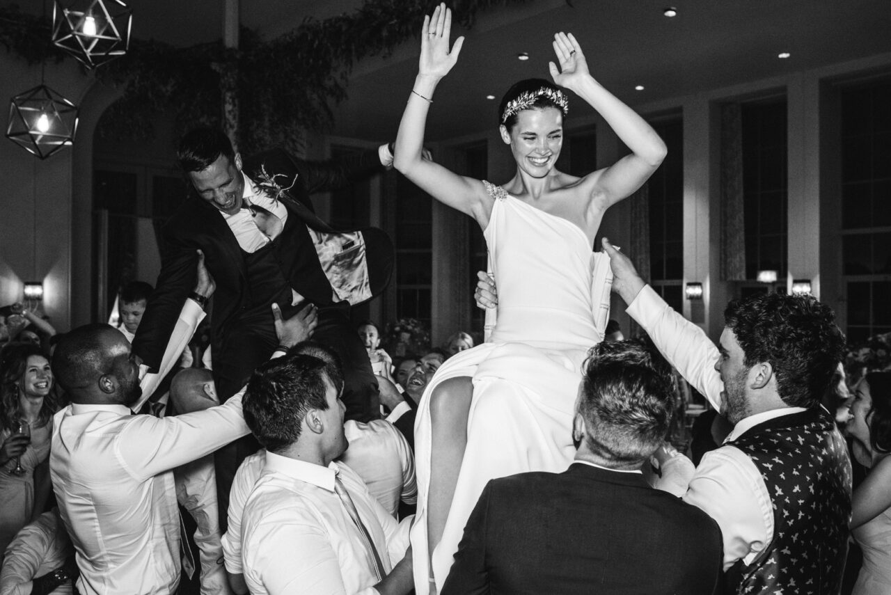 Black-and-white photo of the bride lifted in the air during a lively wedding party