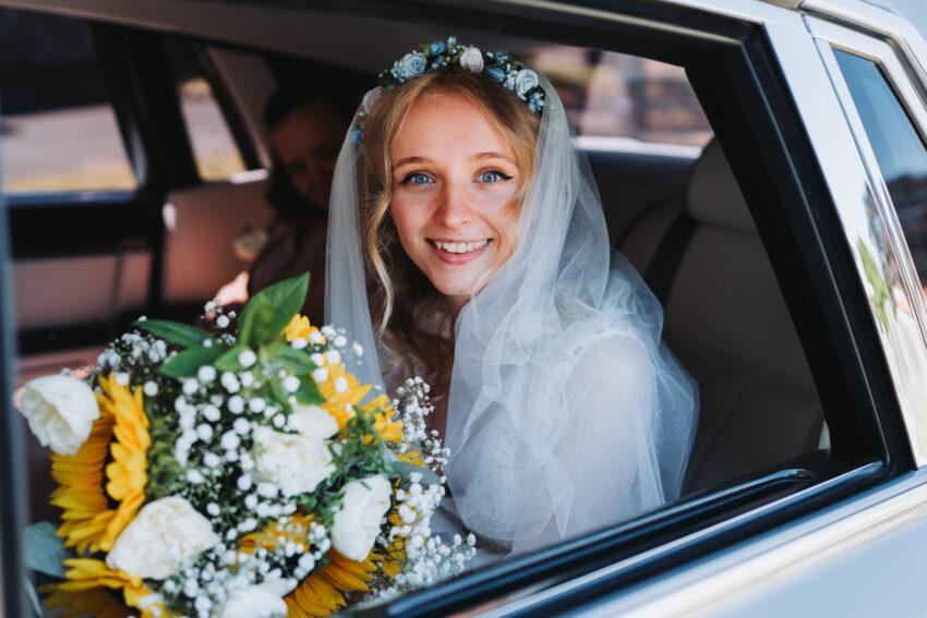 Bride wearing a veil poses with some flowers in the open window of a car.