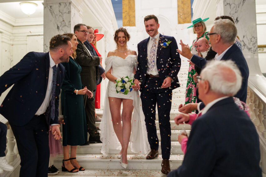 Bride and groom walking down the staircase as guests throw confetti.