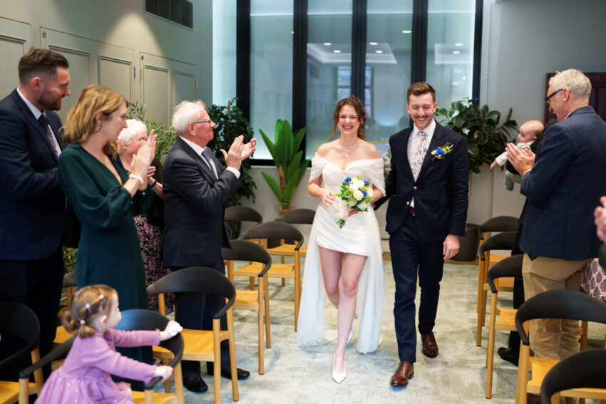 Newly married couple walking down the aisle at Camden Town Hall as guests applaud.