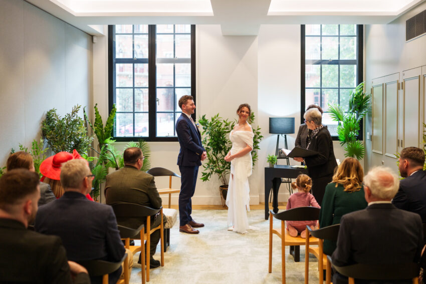 Bride and groom standing side by side during the ceremony at Camden Town Hall, with guests seated behind them.