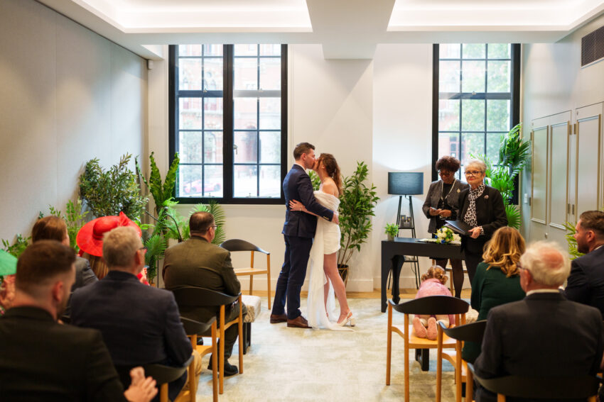 Bride and groom sharing their first kiss during a wedding ceremony at Camden Town Hall, photographed in the modern ceremony room with guests seated around them.