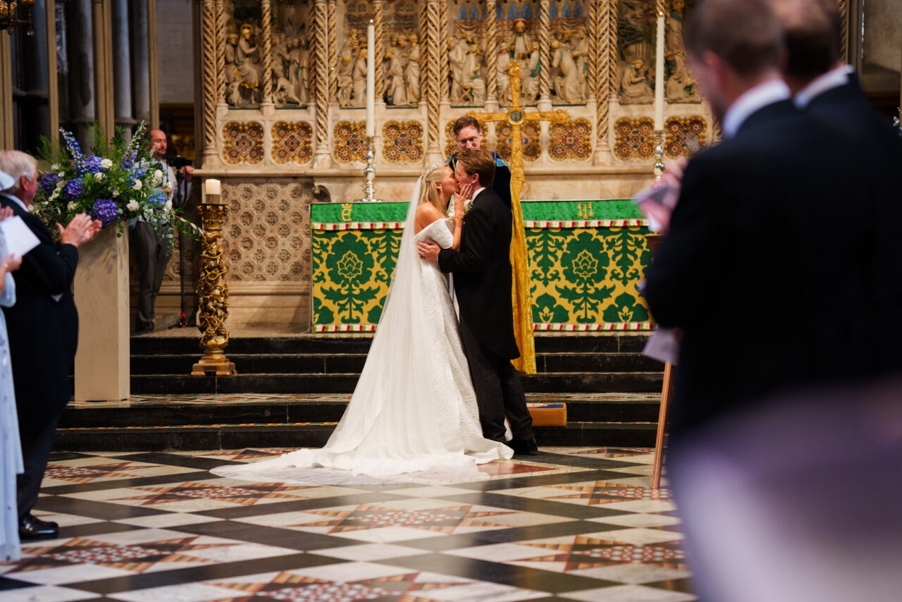 The couple kiss at the altar following the ceremony, guests applauding within a historic Ely church interior.