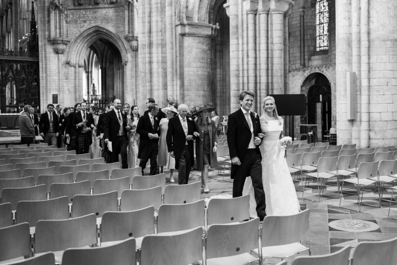Black and white photograph of the couple walking through rows of chairs inside a large historic church, strong vertical columns emphasising scale and perspective.