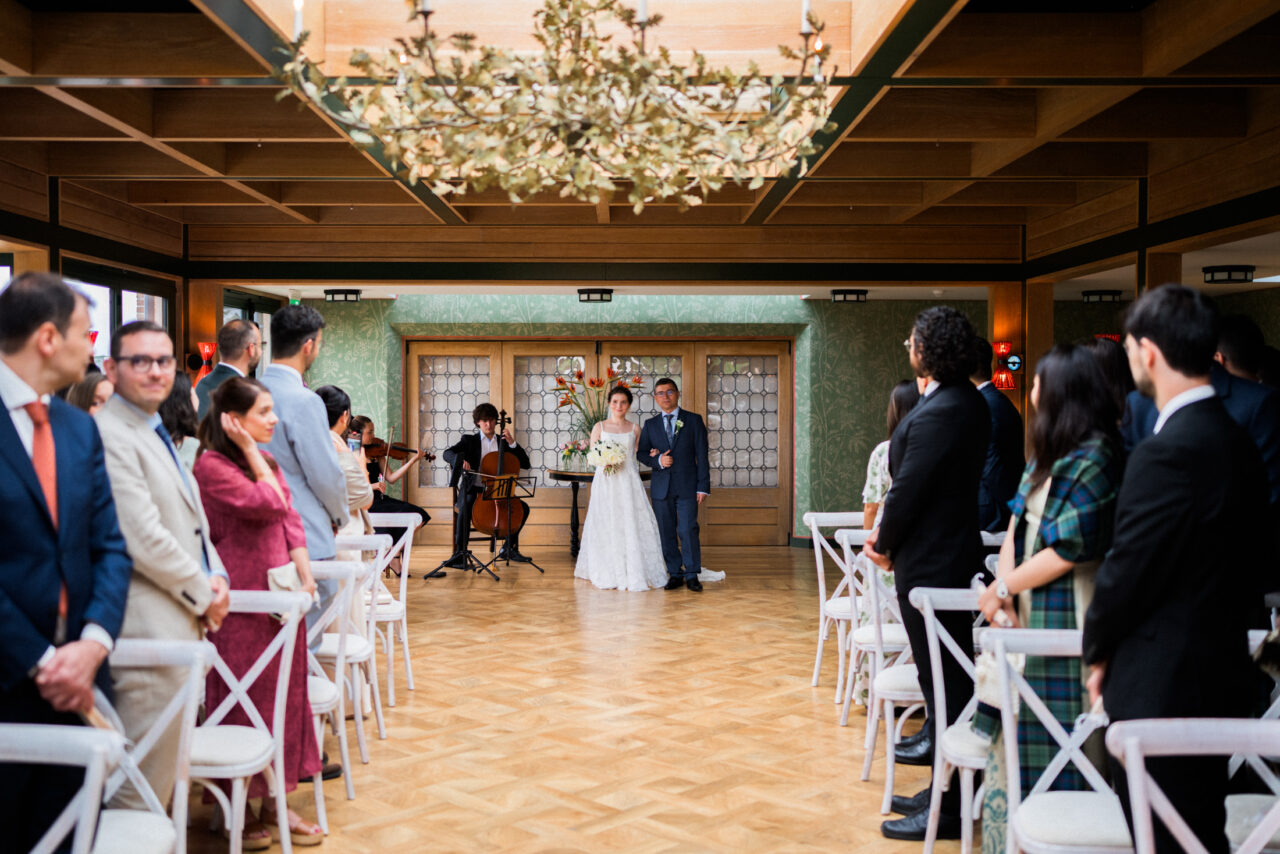Indoor wedding ceremony at The Old Hall in Ely, guests seated on either side, warm interior lighting and wooden floors creating a calm, balanced composition, photographed by an Ely wedding photographer.
