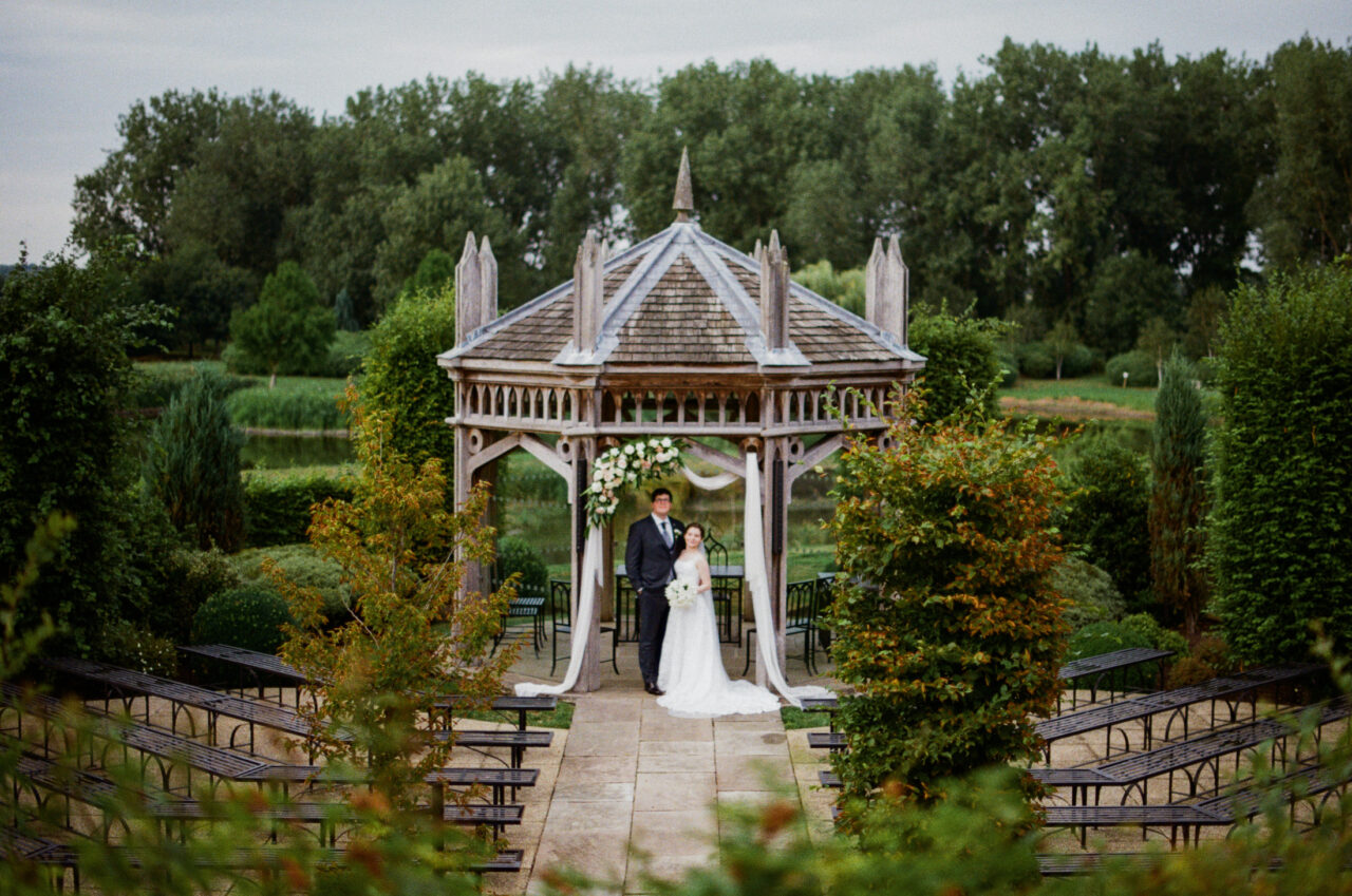 Outdoor ceremony beneath a wooden pavilion, structured garden planting and overcast light giving a soft, even tone to the scene.