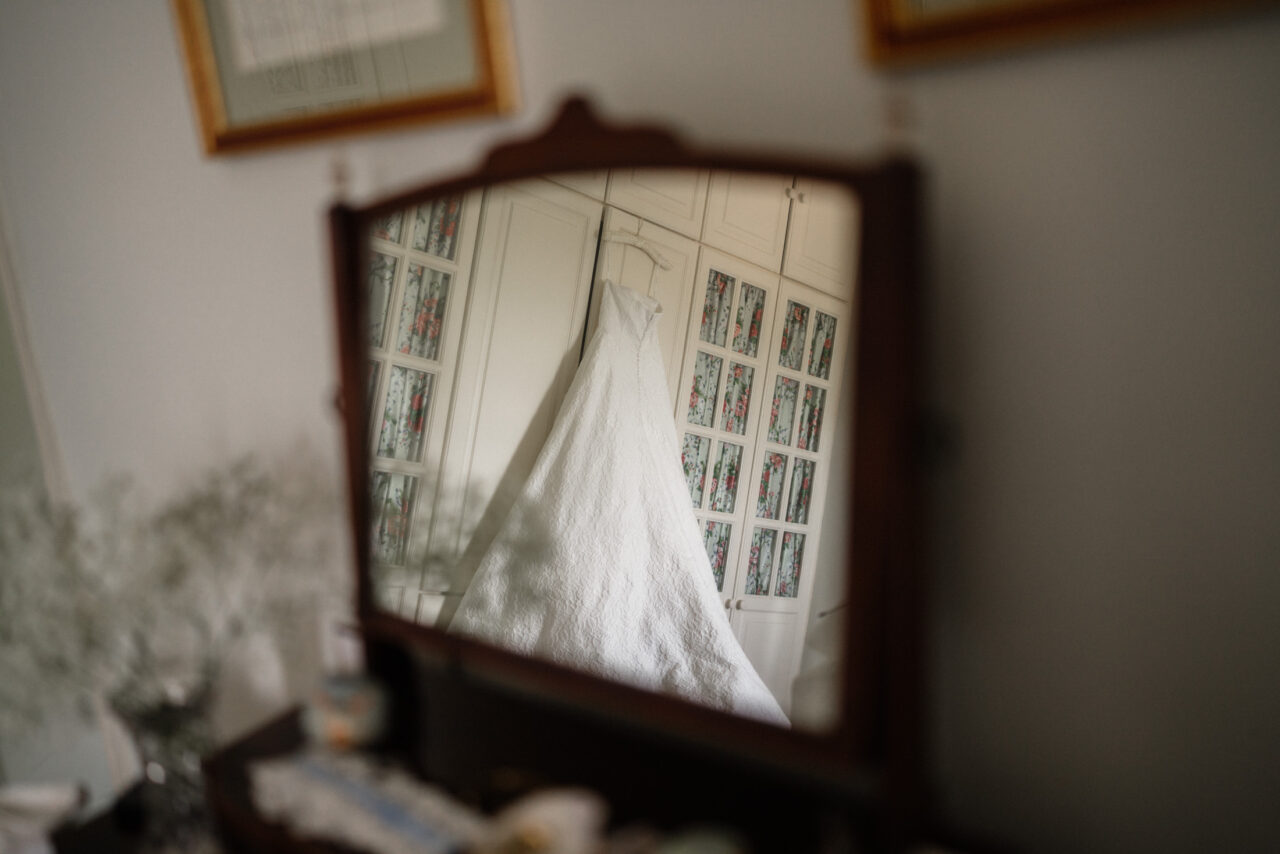 Wedding dress reflected in a mirror during bridal preparations, natural window light and a calm, uncluttered setting.