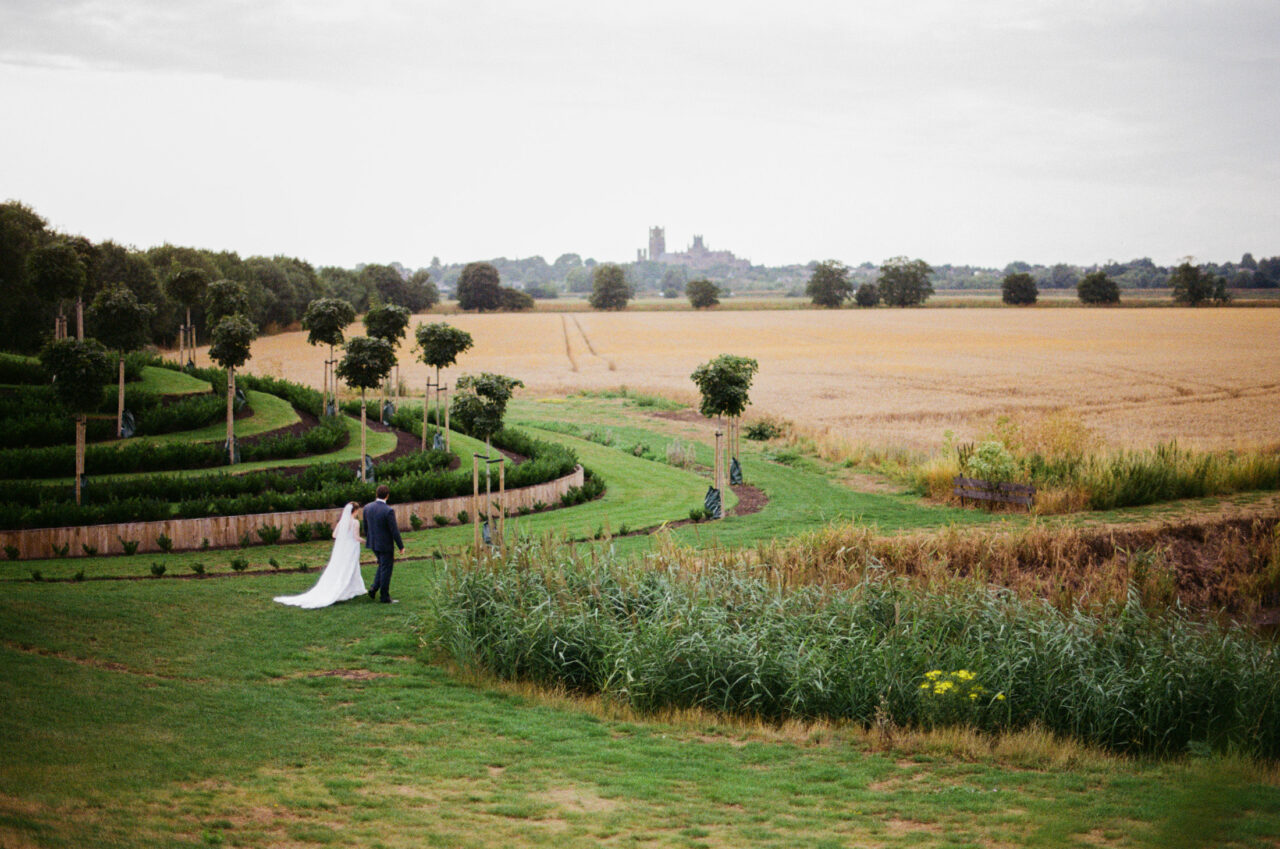 Wide landscape view of the couple walking through formal gardens, winding paths and distant countryside - Ely Cathedral is in the distance.