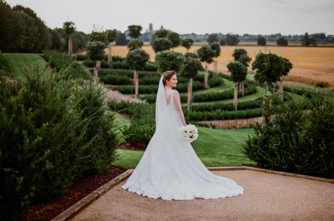 Bride standing alone on a garden path, flowing dress and clipped greenery forming clean lines and gentle contrast.