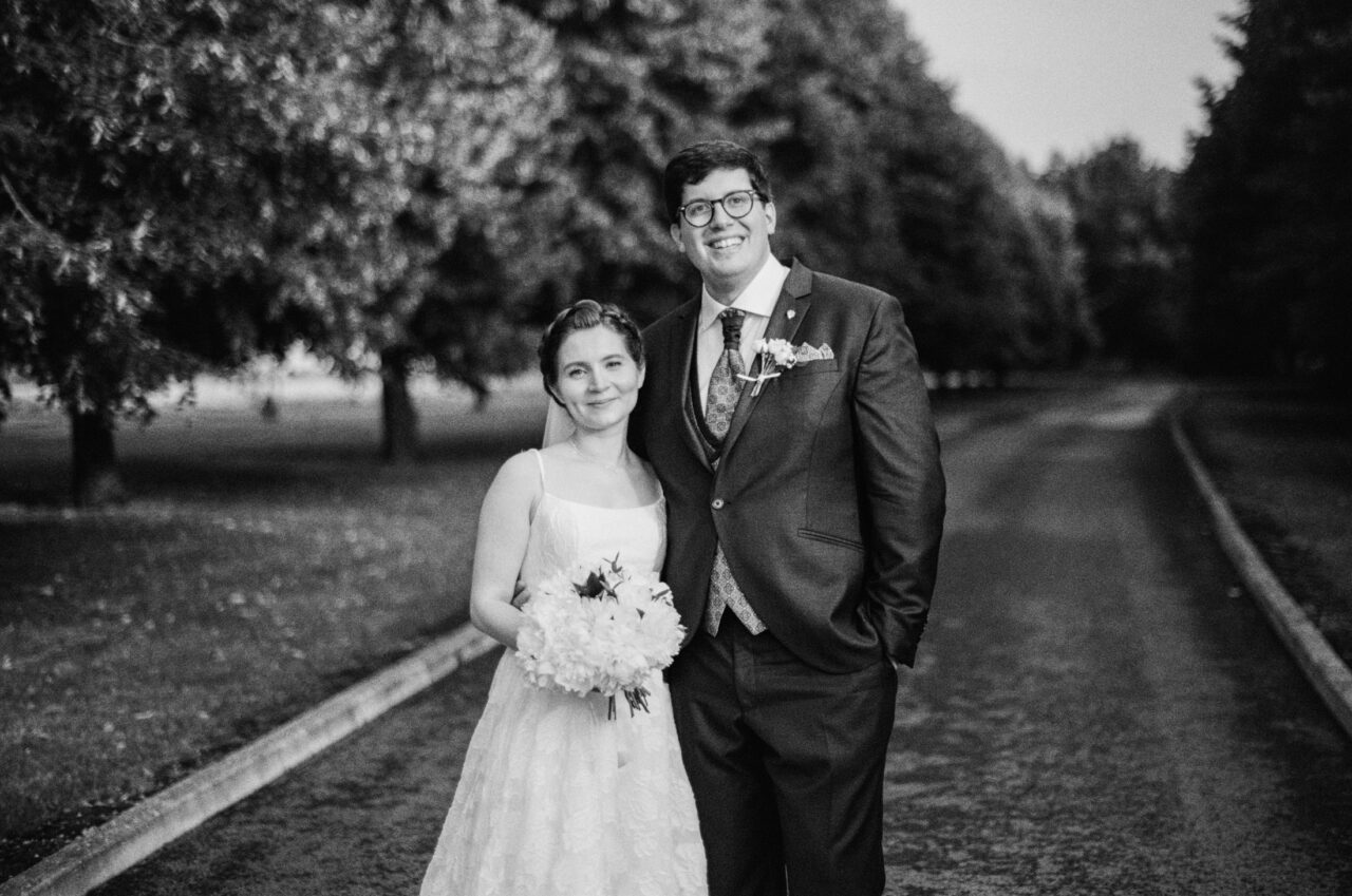 Black and white portrait of the couple walking along a tree-lined path at The Old Hall in Ely, soft light and tonal contrast simplifying the scene.