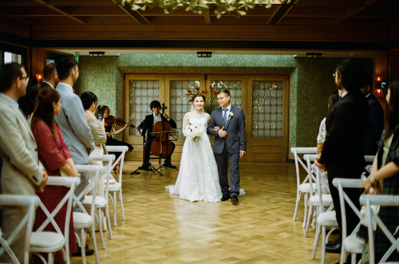 Bride entering the ceremony space with guests seated on either side, warm interior tones and repeating chairs adding structure.