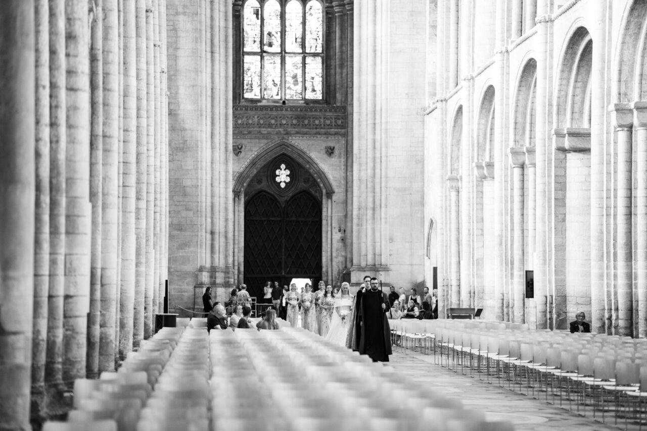 Black and white photograph of guests seated beneath tall stone columns inside a large cathedral-style church, emphasising scale and structure.