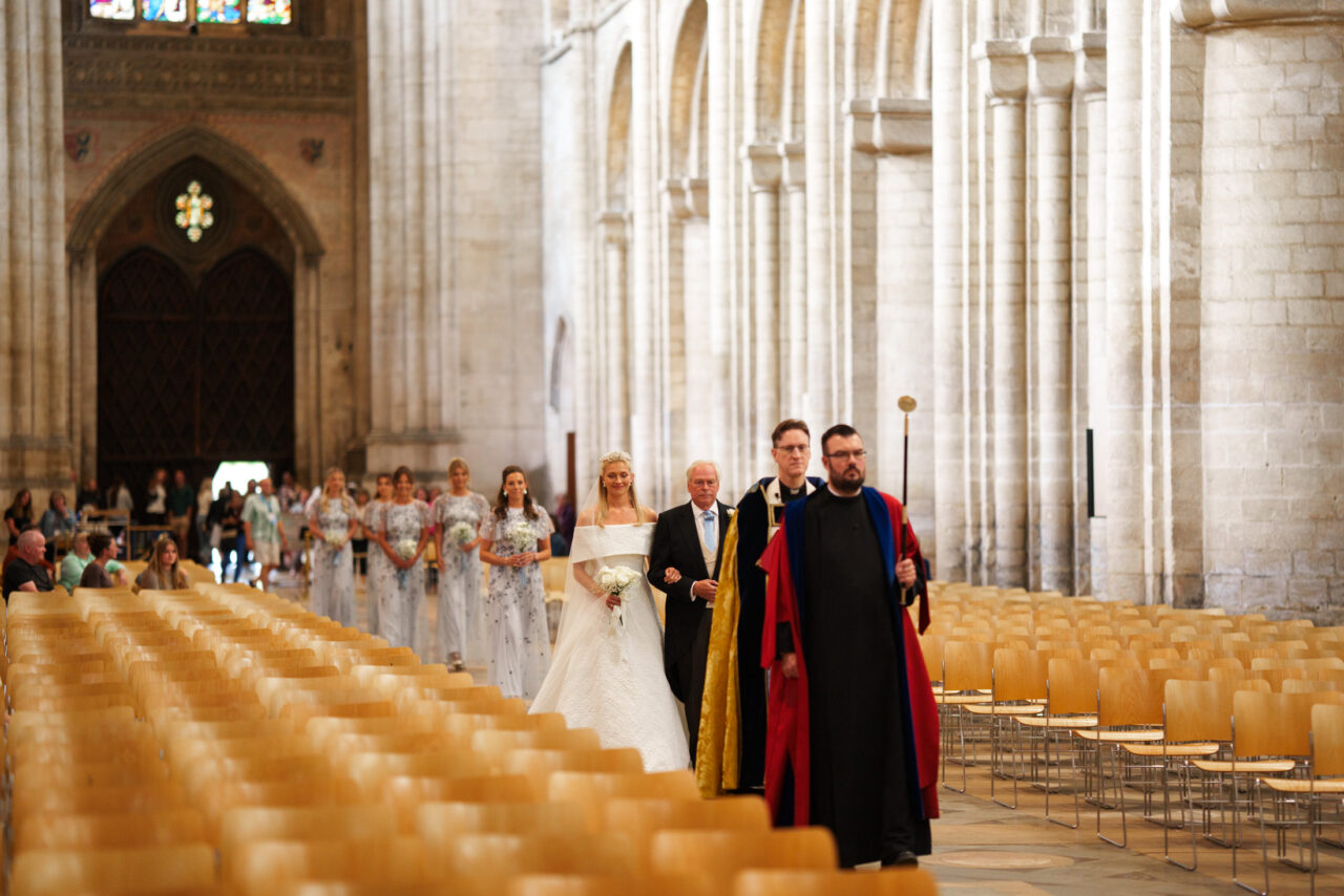 Bridal party beginning their walk down the aisle, repeating chairs and pale stone walls creating strong leading lines.