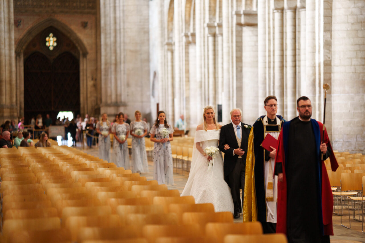 Bride walking down the aisle with her father, soft directional light and shallow focus separating figures from the background.