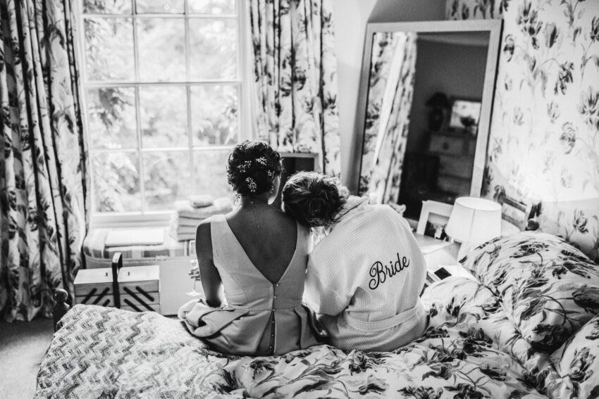 Bride and bridesmaid sitting together on a bed, quiet preparation moment.