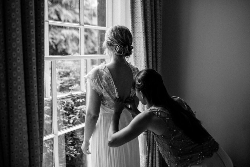Bridesmaid adjusting the back of a wedding dress by a window.