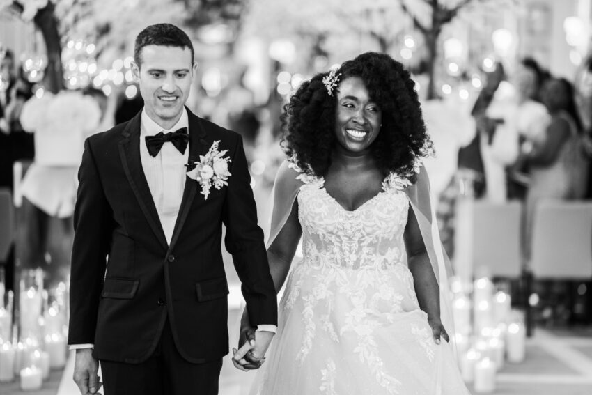 Bride and groom walking hand in hand down a candle-lit aisle at Kimpton Fitzroy London, smiling as they leave the ceremony with soft lights and guests blurred in the background.