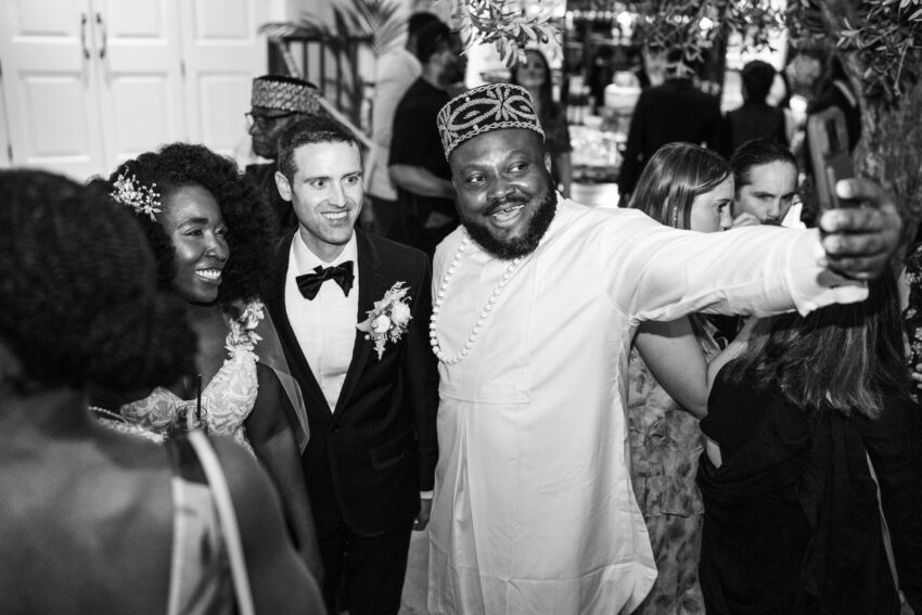 Black and white photograph of the bride and groom smiling among guests during the evening reception, surrounded by raised hands and celebratory gestures.