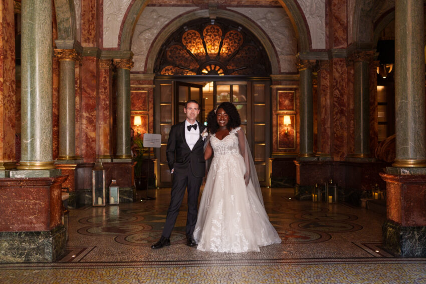 Bride and groom standing together beneath the ornate arched entrance of Kimpton Fitzroy London, with patterned stonework and warm interior lighting behind them.