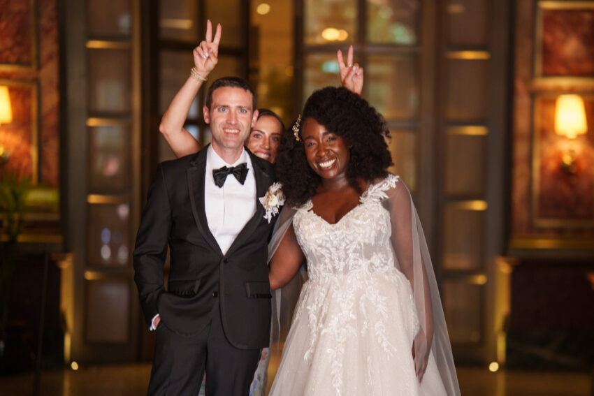 3. Bride and groom posing closely together indoors, smiling as a guest behind them playfully makes bunny ears during the evening celebrations at Kimpton Fitzroy.