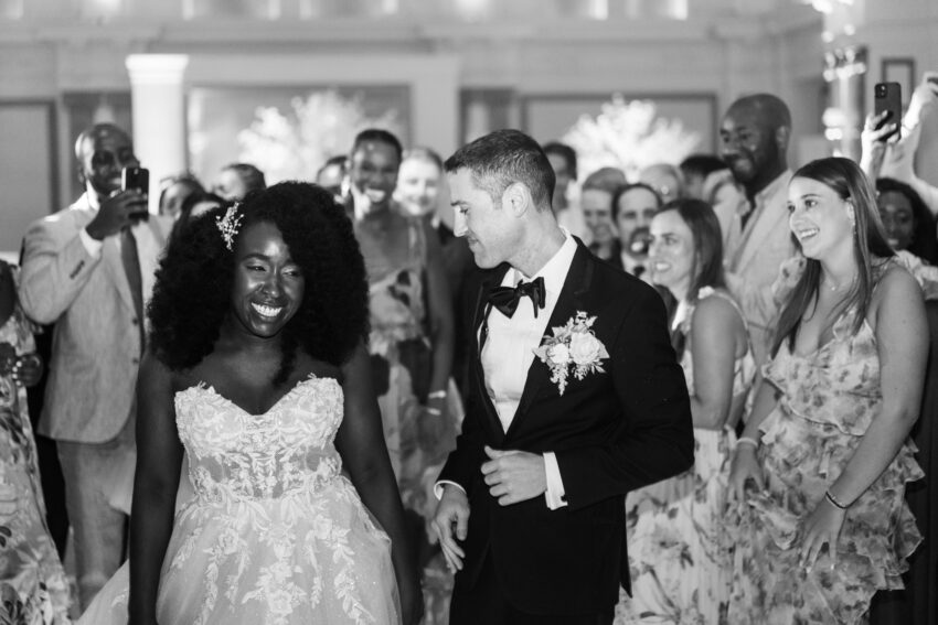 Black and white image of the bride and groom walking through a crowd of guests, smiling as people gather closely around them.