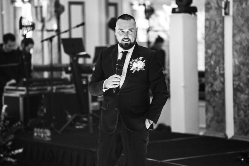 Black and white portrait of a wedding guest holding a microphone mid-speech, standing near the stage area during the evening reception.
